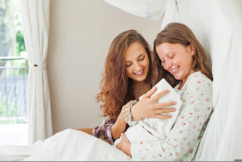 två kvinnor som sitter på sängen ler och ser baby