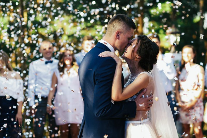 Lyckliga nygifta kysser på verandan under regnet av vita konfetti