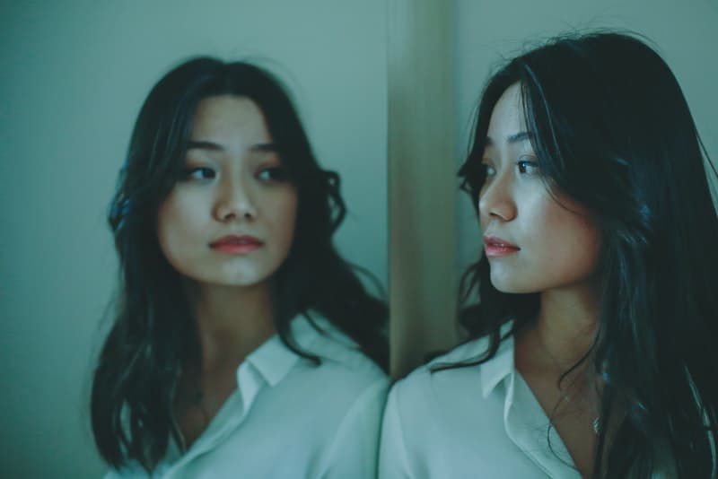 en sorglig kinesisk kvinna i en vit skjorta tittar på sig själv i spegeln