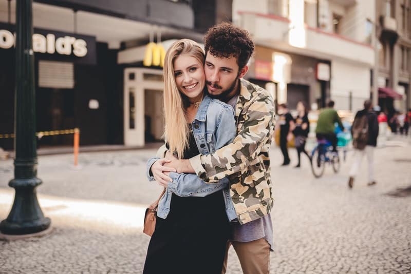 Ein Mann mit Bart hinter dem R&uuml;cken umarmt seine l&auml;chelnde Frau auf der Stra&szlig;e